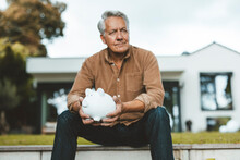 Senior Man With White Hair Holding Piggy Bank At Backyard