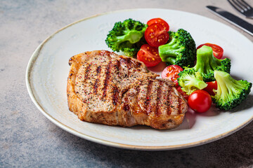 Canvas Print - Grilled pork steak with broccoli and tomatoes on plate, dark background.