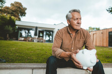 Thoughtful Smiling Senior Man With Piggy Bank Sitting At Backyard