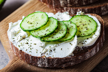 Canvas Print - Toasts with cream cheese, cucumber and rye bread on wooden board, dark background.