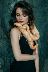 Wall Mural - Closeup portrait beautiful charming modern woman. Closed eyes. Yellow snake on neck, shoulders. Natural makeup, red lips. Brunette girl, long wavy hair, green marble wall. Black elegant evening dress