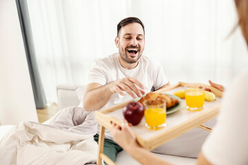 Wall Mural - A happy man having breakfast in the bed brought by his wife at their cozy home.