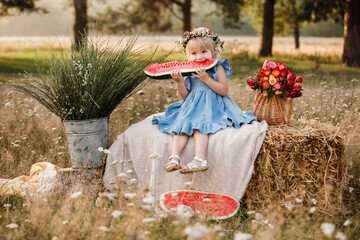 Wall Mural - picnic with family. cute little girl eating big piece of watermelon on straw stack in summertime in the park. Adorable child wearing in flowers wreath on head and blue dress.