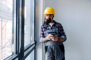 A happy, bearded worker with a helmet on his head, is standing next to a window and using his phone to check when is material arriving. He is looking through the window.