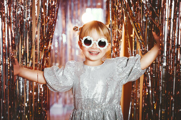 Happy little stylish girl in shiny dress having fun. Festive background with foil curtain decorations for kids birthday or fancy dress party, disco music or New Year.