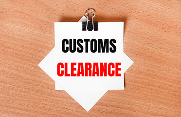 On a wooden table under a black paper clip lies a sheet of white paper with the text CUSTOMS CLEARANCE