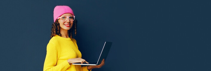 Portrait of stylish modern young woman working with laptop wearing colorful clothes on dark blue background, banner blank copy space for advertising text