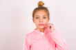 caucasian little kid girl with bun hairstyle wearing pink tracksuit over white background mouth and lips shut as zip with fingers. Secret and silent, taboo talking.