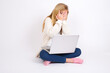 © Jihan - caucasian teen girl sitting with laptop in lotus position on white background covering face with hands and peering out with one eye between fingers. Scared from something or someone.