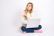 © Jihan - Portrait of caucasian teen girl sitting with laptop in lotus position on white background posing on camera with tricky look, presenting product with index finger. Advertisement concept.