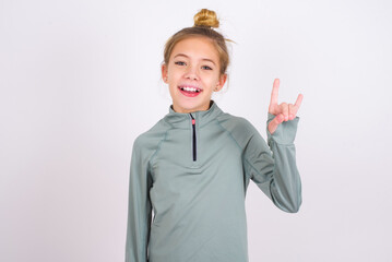 Wall Mural - little caucasian kid girl with hair bun wearing technical shirt over white background doing a rock gesture and smiling to the camera. Ready to go to her favorite band concert.