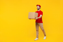 Full Body Side View Happy Delivery Guy Employee Man In Red Cap T-shirt Uniform Workwear Work As Dealer Courier Hold Cardboard Box Isolated On Plain Yellow Background Studio Portrait. Service Concept.