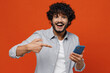 © ViDi Studio - Jubilant exultant happy young bearded Indian man 20s years old wears blue shirt hold in hand use mobile cell phone pointing index finger on screen isolated on plain orange background studio portrait.