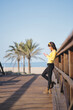 © Fotografia Juan Reig - Vertical photo of a woman leaning on the railing of a bridge next to the beach while using a mobile