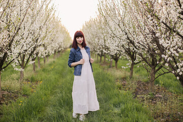 Full length portrait of charming young woman wearing white dress and denim jacket spending leisure time among beautiful blooming garden. Concept of people, beauty and relaxation.