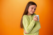 Sleepy teenager girl holding a hot cup of coffee on orange background