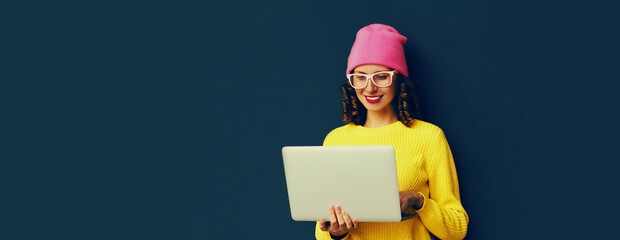 portrait of stylish modern young woman working with laptop wearing colorful clothes on dark blue bac