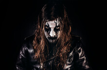Portrait Photo Of Black Metal Metalhead Man With Long Brown Hair And Painted Face Standing In Leather Jacket And Looking At Camera On Black Background.