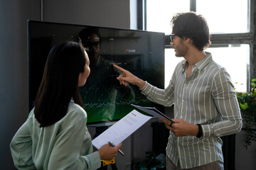 Aufkleber - Young confident male economist pointing at graph on interactive screen during presentation of financial data to Asian female colleague