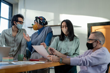 Aufkleber - Young Asian female economist and mature businessman in protective masks discussing paper against two communicating coworkers