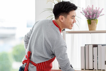 Asian happy male husband housekeeper servant in maid uniform with rubber gloves and red apron standing smiling posing holding feather duster whisker in hands cleaning wiping living room at home