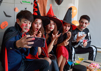 Wall Mural - Group of Asian young scary creepy male and female friends in Halloween witch sorcerer ghost and skeleton costume sitting on sofa taking selfie photo together in traditional trick or treat night party