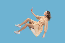 Hovering In Air. Smiling Girl In Yellow Dress Levitating With Mobile Phone, Reading Message Chatting Happy Joyful In Social Network Online, Surfing Web While Flying. Indoor Shot Isolated On Blue