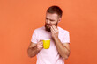 Portrait of unhealthy young adult bearded man having teeth pain after drinking hot or cold beverage, dental injury, wearing pink T-shirt. Indoor studio shot isolated on orange background.