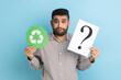 © khosrork - Portrait of bearded doubtful confused businessman standing holding green recycling sign and paper with question mark, wearing striped shirt. Indoor studio shot isolated on blue background.
