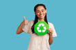 © khosrork - Positive woman with black dreadlocks looking at camera with toothy smile, showing thumb up, holding recycling sign, wearing white shirt. Indoor studio shot isolated on blue background.
