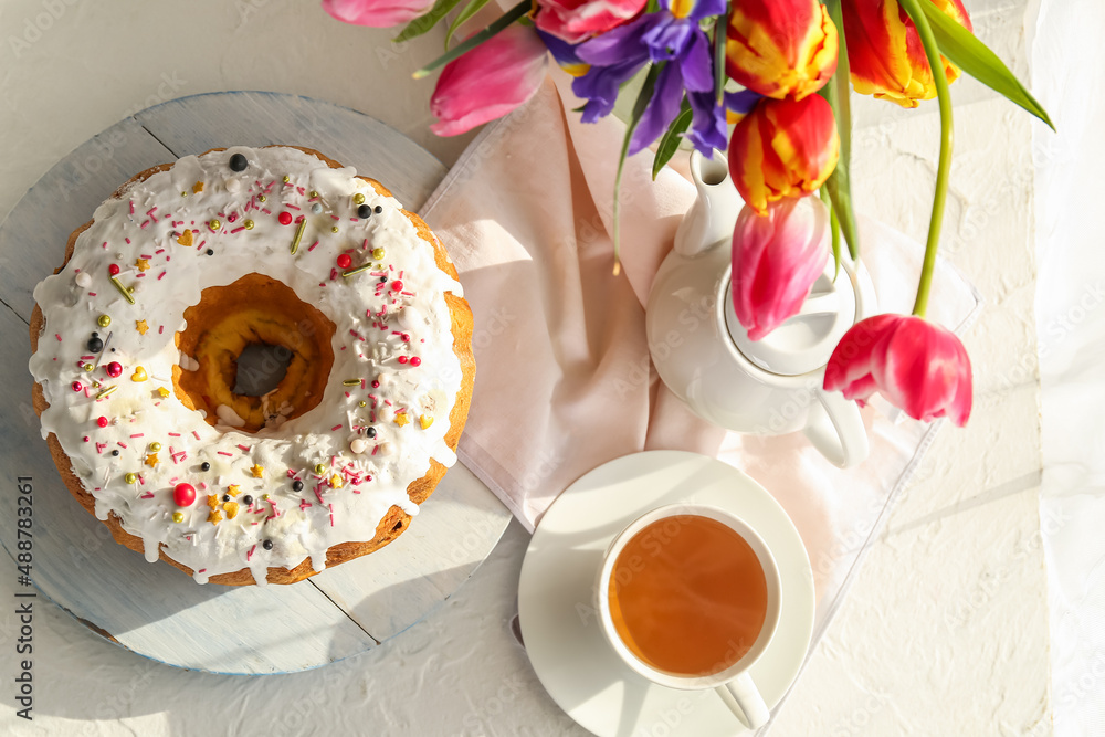 Tasty Easter cake, cup of tea and vase with flowers on light background