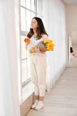 Wall Mural - Pretty young Asian woman with bouquet of flowers drinking tea near window