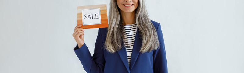 Wall Mural - Pretty middle aged Asian lady with long grey hair wearing blue jacket holds card with word Sale posing on light background in studio