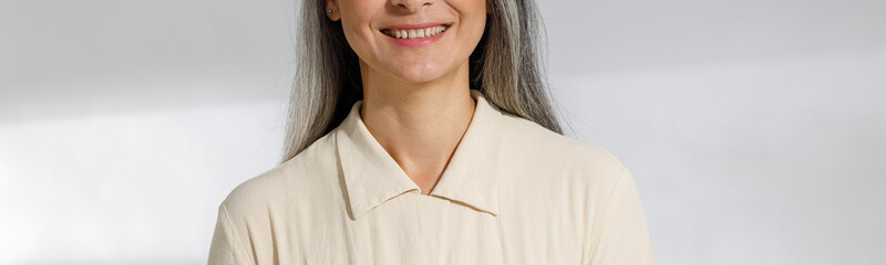 Wall Mural - Portrait of pretty smiling grey haired woman in beige blouse standing on light background in studio. Mature beauty lifestyle