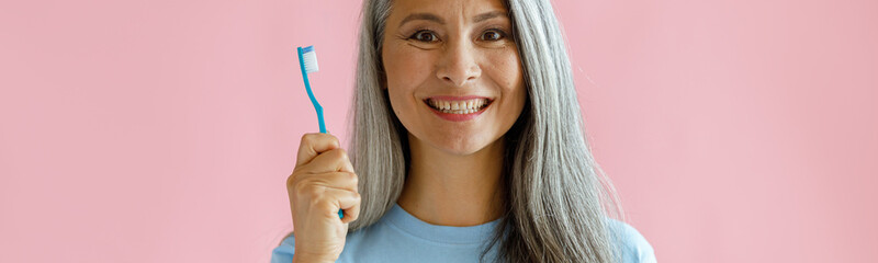 Wall Mural - Cheerful middle aged Asian woman with grey hair in blue t-shirt holds toothbrush standing on pink background in studio. Oral cavity hygiene