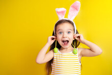 Happy Easter. A Funny Crazy Girl With Bunny Ears Holds Green Eggs In Her Hands As Earrings And Is Surprised And Her Mouth Is Open. Yellow Background. Space For Text