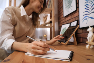 Wall Mural - Young woman using tablet while drawing in sketchbook with pencil at wooden table indoors, focus on hand