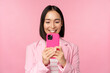 Image of smiling asian corporate woman in suit looking, watching on smartphone app, using mobile phone application, standing over pink background