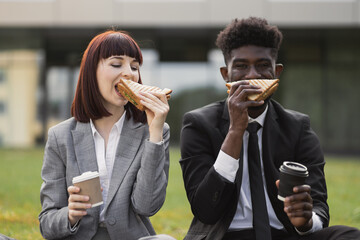 Healthy sandwich, lunch break concept. Two multiracial successful partners sitting on green grass outside modern office, enjoying their lunch with sandwiches and coffee on fresh air