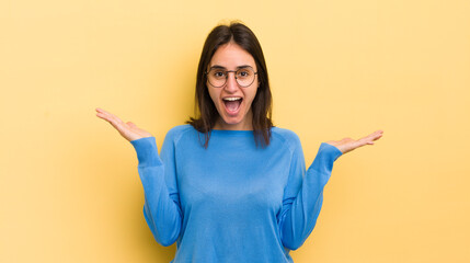 young hispanic woman looking happy and excited, shocked with an unexpected surprise with both hands open next to face