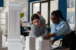 © DC Studio - Architectural engineers doing teamwork reading construction plans over table with white foam scale model of buildings. Team of two architects colleagues collaborating looking down at blueprints.
