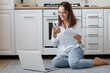 © sementsova321 - Portrait of smiling satisfied woman wearing white shirt and jeans sitting on floor, holding mobile phone, looking at pc computer with positive facial expression.