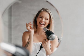 Wall Mural - Wash hair, care and dry. Playful young lady doing hair styling with hair dryer and having fun, standing near mirror