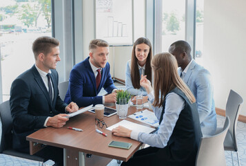 Wall Mural - Young handsome man gesturing and discussing something while his coworkers listening to him sitting at the office table.