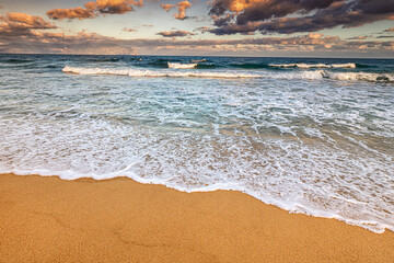 Wall Mural - Wide angle view of an idyllic sea sandy beach with an incoming azure wave at high tide. Windy and stormy weather at sunset. Background for travel and vacation