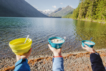 Friends on a hike eat porridge for breakfast in tourist plates and bowls. Against the background of an idyllic landscape of a mountain lake. Food and cooking