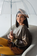 Wall Mural - stylish asian woman sitting in armchair under umbrella and smiling at camera isolated on grey