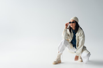 Wall Mural - full length view of asian woman in white clothing adjusting sunglasses and looking away on grey