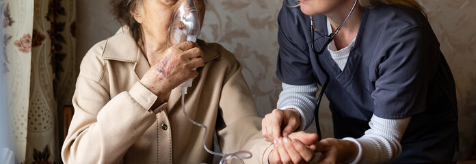 Wall Mural - Young high-skilled pleasant woman doctor putting on nebulizer mask on face of her elderly female patient to make inhalation when visitng him at home. Flu, cold and cough treatment.