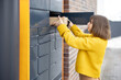 © rh2010 - Young woman getting parcel from cell of automatic post terminal outdoors. Concept of contactless and smart delivery. Idea of modern shipping and logistics. Woman wearing yellow sweater